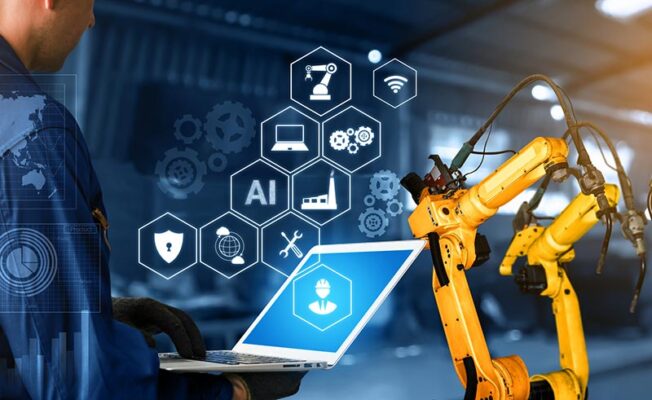 A person uses a laptop in an industrial setting, surrounded by digital icons and yellow robotic arms.