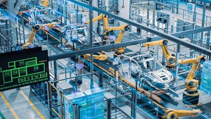 Car frames sit in a row in an automotive manufacturing facility enabled by Industry 4.0 solutions while yellow robot arms hover over them.