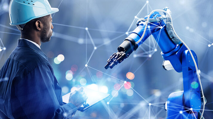 A person in a hard hat operating a tablet and overseeing a robotic arm in an industrial environment enhanced by 5G connectivity.