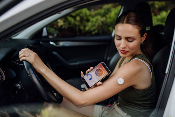 A woman sitting in a car uses her phone to check the glucose sensor on her arm.