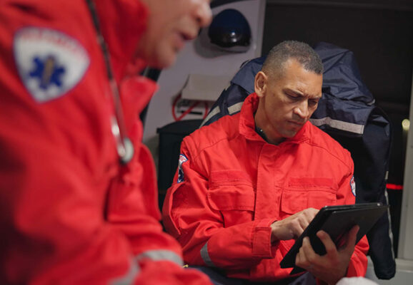 Two first responders in red uniforms are inside a vehicle. One is focused on a tablet, while the other is partially visible in the foreground.