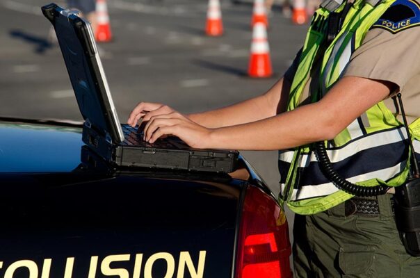 A first responder in a uniform and reflective vest types on a laptop placed on the trunk of a patrol car.