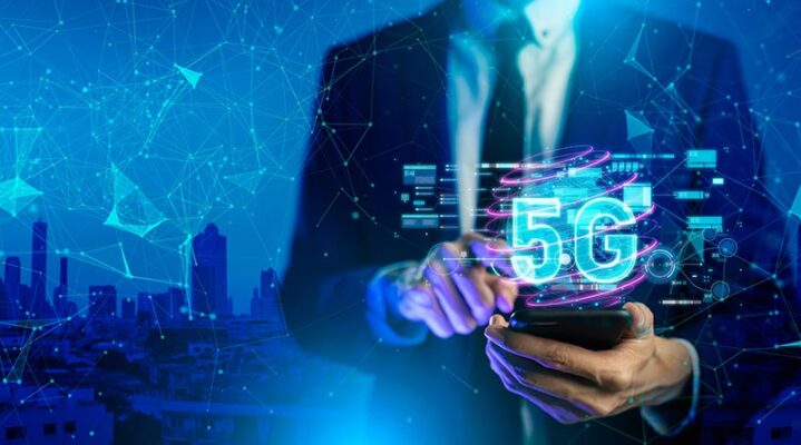 A person in a suit uses a smartphone with a glowing 5G hologram, surrounded by digital network graphics over a cityscape.