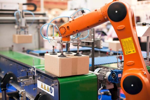A robotic arm with suction cups, powered by private 5G networks, is lifting a cardboard box onto a conveyor belt in an industrial setting.