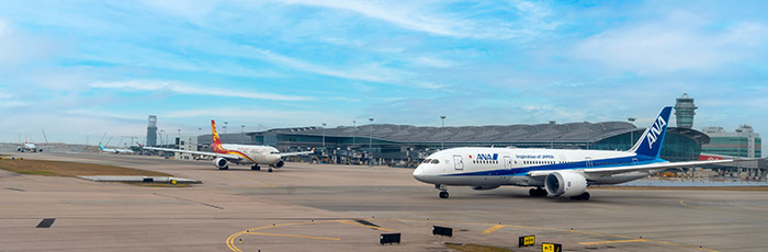 Wide view of an airport runway with two airplanes taxiing and airport terminal buildings in the background, all under a partly cloudy sky.