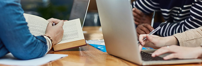 In a quiet corner, individuals sitting at a table are studying, with one person reading an open book and another typing on a laptop.