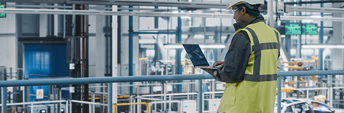 A person wearing a yellow safety vest and helmet works on a laptop connected to a private 5G network on an elevated platform within a large manufacturing facility filled with machinery.