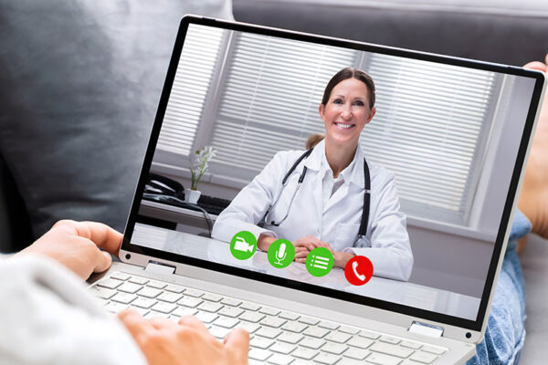 A person uses a laptop to have a virtual consultation with a smiling doctor who is wearing a white coat and a stethoscope.