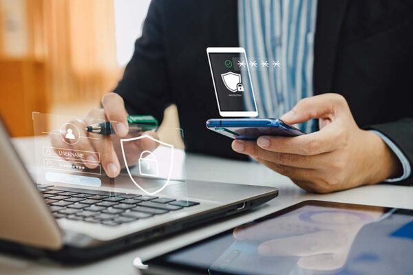 A person uses a smartphone and laptop, surrounded by digital security icons, highlighting an IoT security strategy with multifactor authentication.