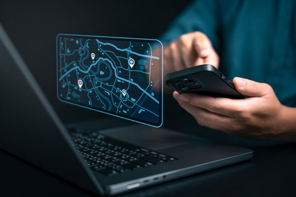 A digital map interface with location pins hovers over a laptop keyboard while hands in the background type on a cell phone. 