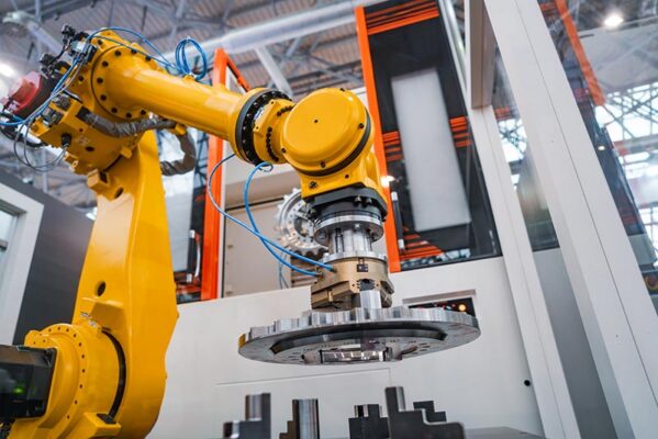 A yellow industrial robotic arm is assembling or inspecting a large metal component in a factory or manufacturing setting.