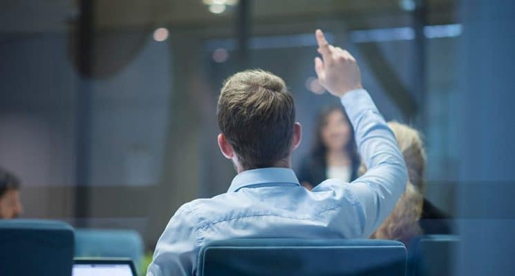 A person with their hand raised to ask a question. Asking the right questions will help you choose the best IoT module provider.