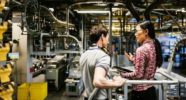 Two people standing in a manufacturing plant, discussing the information they are viewing on the tablet. The manufacturing industry has adopted IoT technology to capture data to improve decision-making and streamline operations.