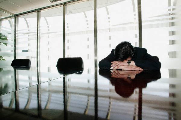 A disappointed person alone in a conference room resting their head on their hands, which are crossed on a glossy black table.