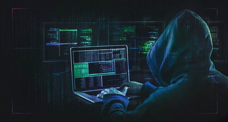 Hooded figure in a dark room working on a laptop, representing a hacker.