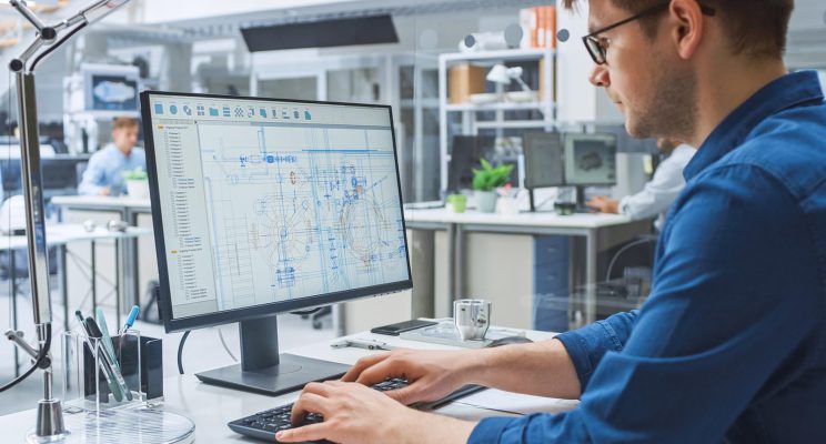 An engineer working on a computer in an office with other engineers. The monitor shows technical drafts and drawings.