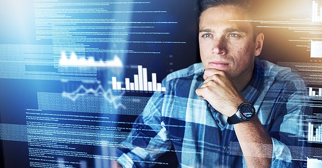 A person sitting thoughtfully in front of a monitor that's showing a stream of data and graphs.