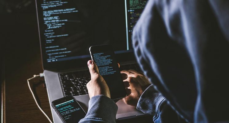 A hooded figure holding a cell phone in one hand and typing on a computer with the other. Another cell phone is sitting on the laptop. The screens show backend-style data to represent hacking.