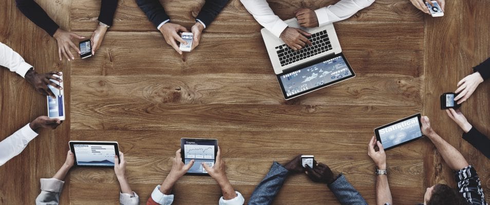 Overhead view of people sitting at a conference table, using smartphones, tablets and laptops.