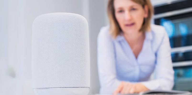 A woman is looking at a smart speaker in front of a laptop.