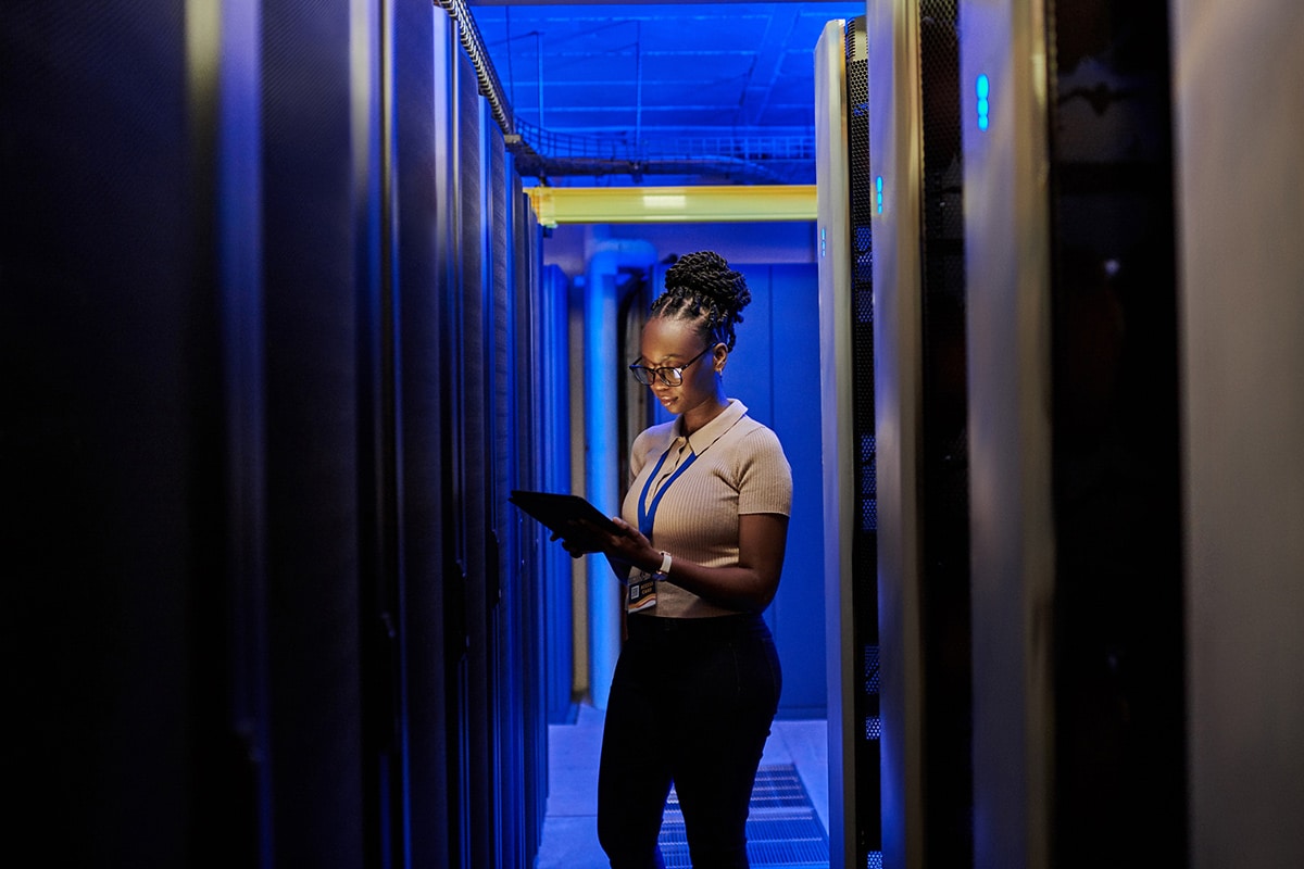 A woman standing in a server room, holding a tablet to monitor the IoT AppZone.