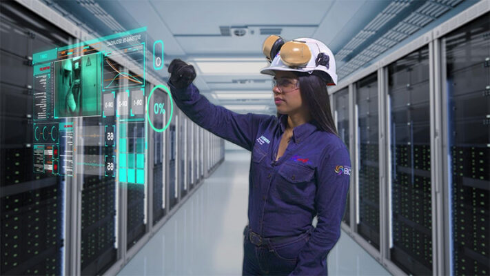 A woman in protective gear interacts with a transparent digital interface in a server room.