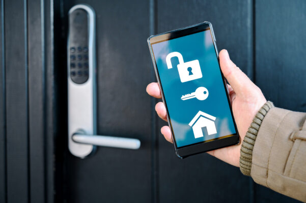 A hand holds a smartphone displaying lock, key and house icons on a blue background next to a digital keypad door lock.