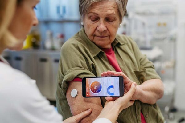 A health care professional views a digital glucose reading on a cell phone held in front of an older woman with a glucose monitor attached to her upper arm.