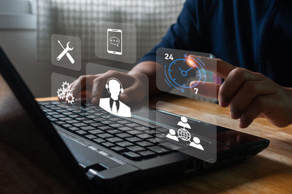 A person taps one of several virtual icons above a laptop keyboard. The icons show 24/7 support, customer service and global communication symbols.
