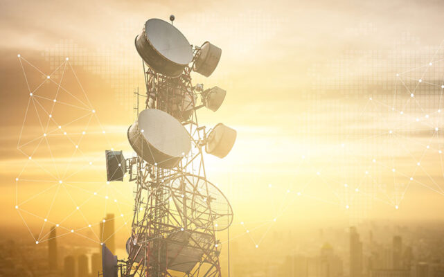 A telecommunications tower with multiple satellite dishes and antennas against sunset-lit cityscape in the background.