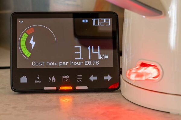 A digital electricity smart meter displays 3.14 kW usage at a cost of £0.76 per hour. A plugged-in appliance with a red light sits nearby.