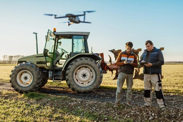 Two people stand by a tractor in a field on a clear day, operating a drone with remote controls.