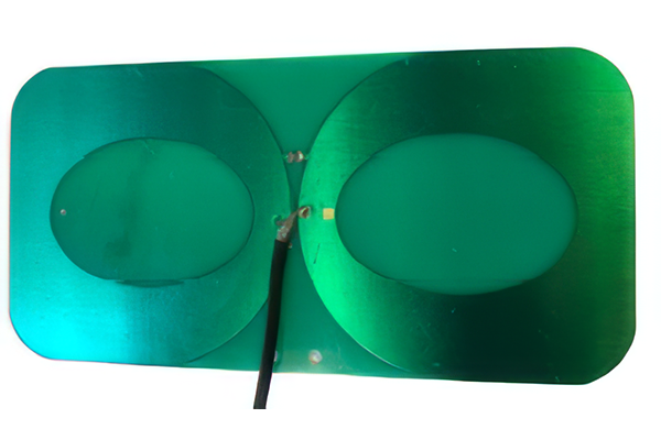 A green rectangular circuit board with two large overlapping oval-shaped copper coils and a black wire attached at the center.