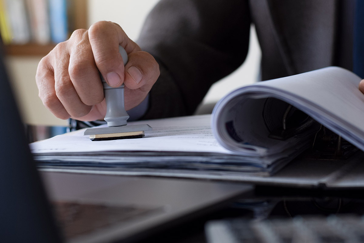 A man in a suit is approving a document with a rubber stamp for IoT device compliance.