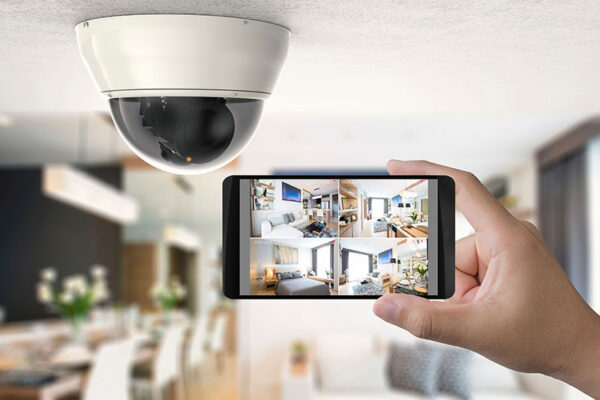 A hand holds a smartphone displaying live security camera feeds in a modern home, showcasing critical IoT solutions at work, with a dome security camera mounted on the ceiling in the background.