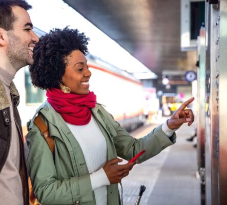 Two people smiling at a train station, one pointing at a smart vending machine.