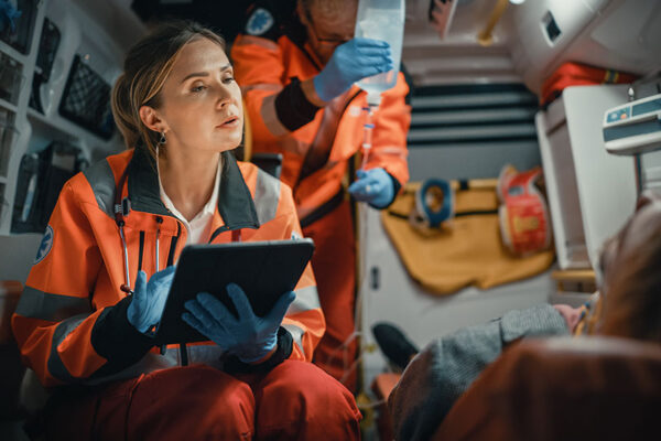 A paramedic is in an ambulance beside a patient. The paramedic is using a tablet, showing an example of 5G in health care during transport.