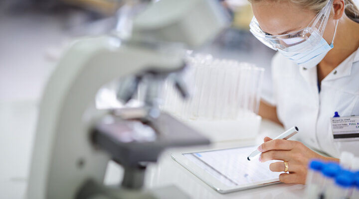 A person wearing safety goggles and a mask works on an electronic tablet beside a microscope in a laboratory.