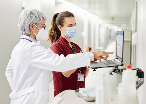 Two medical professionals dressed in masks and scrubs review information on a laptop in a clinical setting.
