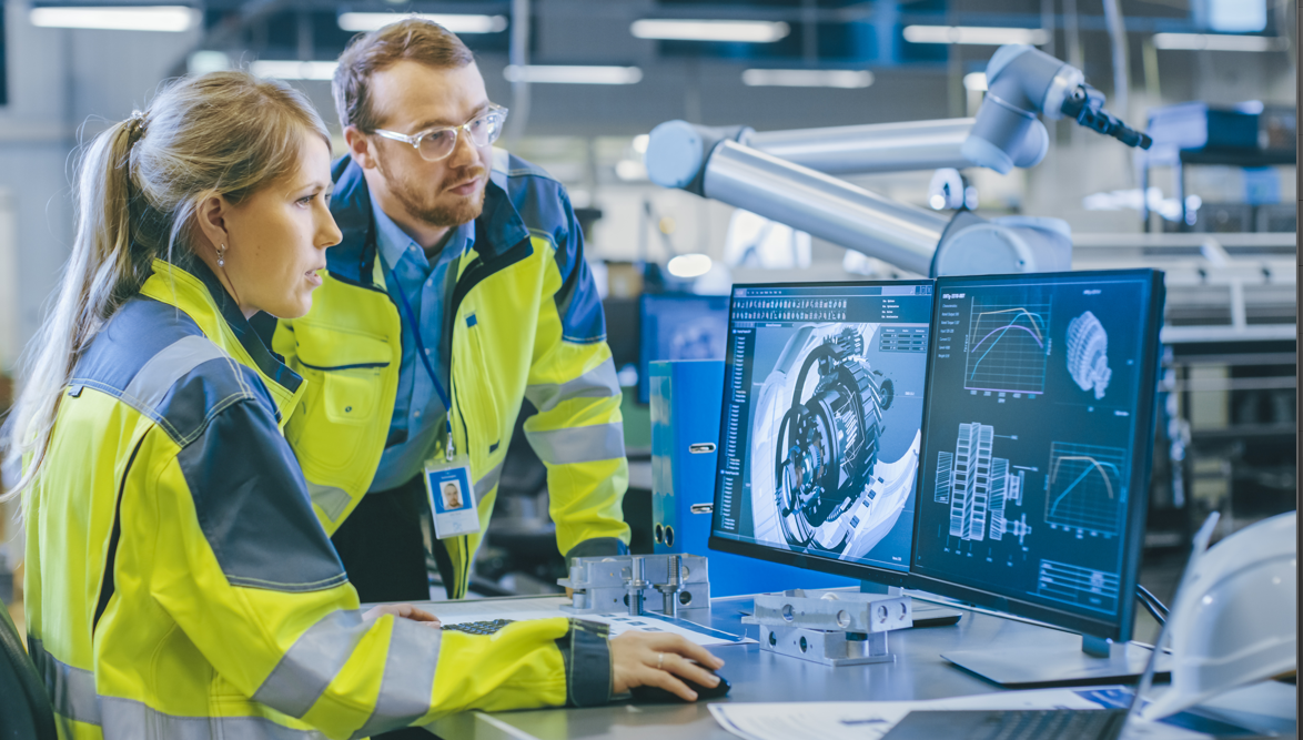 Two engineers in high-visibility vests discuss 3D machine part models on a computer screen in an industrial setting.