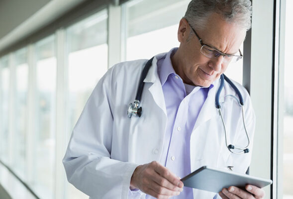 A doctor wearing a white coat and stethoscope looks at a tablet device.