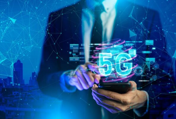 A businessperson holding a smartphone that displays a holographic 5G symbol, with a futuristic cityscape and IoT cybersecurity graphics in the background.