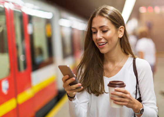 A young person smiles while looking at their smartphone with a red train in the background.
