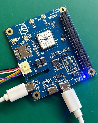 A blue circuit board with various electronic components, including a microcontroller, resistors, capacitors, and connectors. Two white cables are plugged into the board.