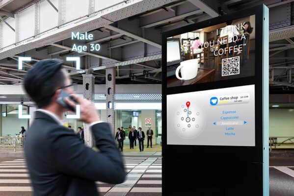 A person in a suit talks on a phone and stands in front of a digital signage display showing a coffee-related advertisement and a menu with different coffee options at a train station.