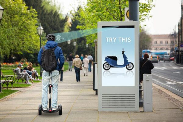 A person riding a Segway looks at a digital advertisement board displaying a blue scooter with the text "TRY THIS" in an urban park setting.