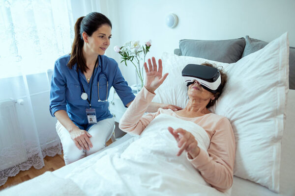 A woman lying in bed wears a virtual reality headset, experiencing a medtech innovation, with a health care worker seated nearby.