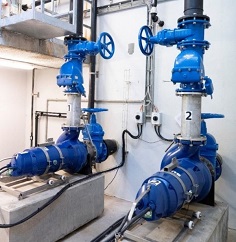 Two large blue industrial valves connected to pipes in a clean mechanical room. The valves are labeled with numbers and the surrounding area features electrical wiring and a concrete floor.