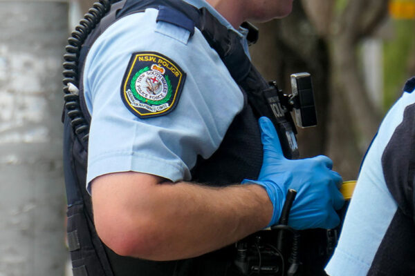 A police officer wearing a blue uniform and gloves, equipped with a radio and body-worn camera.