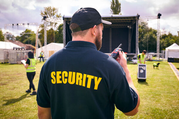 A security guard speaks into a walkie-talkie at an outdoor event, while the stage and crew members bustle in the background.